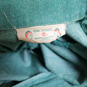 May include: Close-up of a teal green fabric with a white label. The label reads "Girl Scouts" with illustrations of girls and the text "National Equipment Service, New York N.Y.". The fabric has a textured appearance and a button is visible.