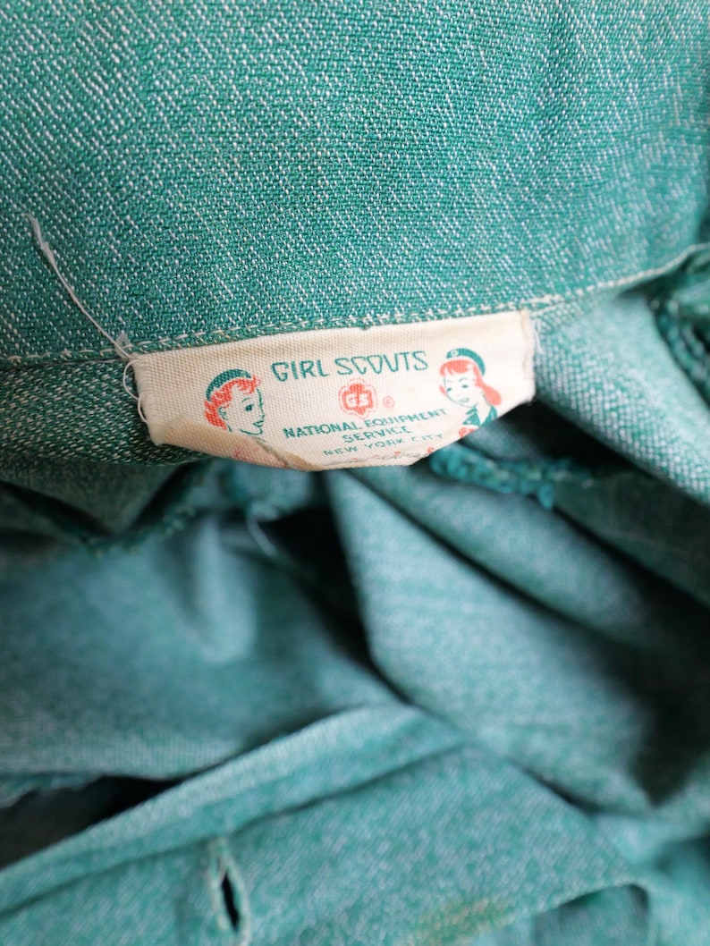 May include: Close-up of a teal green fabric with a white label. The label reads "Girl Scouts" with illustrations of girls and the text "National Equipment Service, New York N.Y.". The fabric has a textured appearance and a button is visible.
