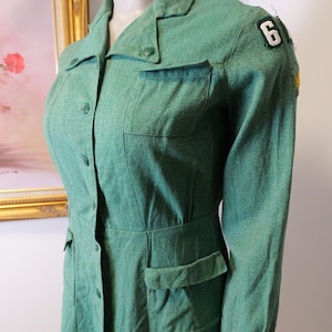 May include: A vintage green dress with long sleeves, a collared neckline, and button-down front. The dress features two chest pockets, two hip pockets, and a patch with the number 6 on the shoulder. The dress is displayed on a mannequin.