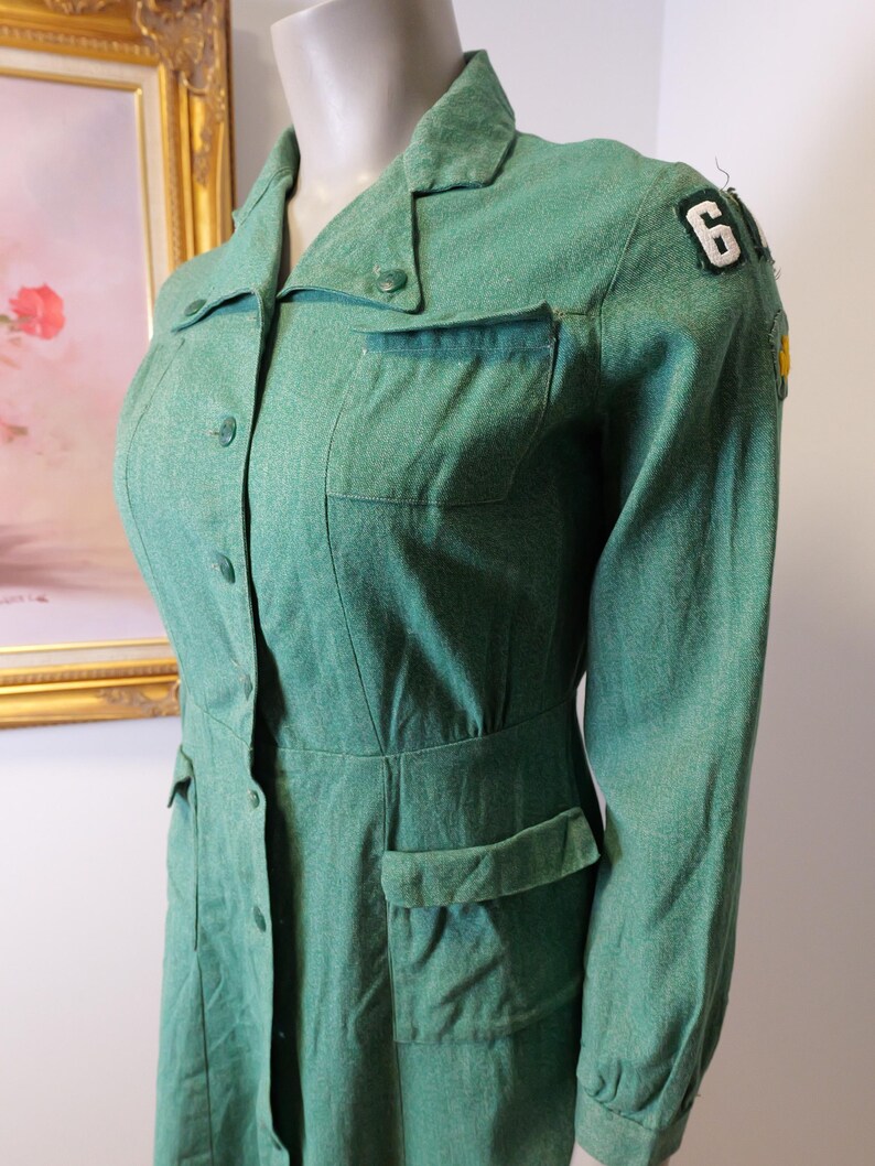 May include: A vintage green dress with long sleeves, a collared neckline, and button-down front. The dress features two chest pockets, two hip pockets, and a patch with the number 6 on the shoulder. The dress is displayed on a mannequin.