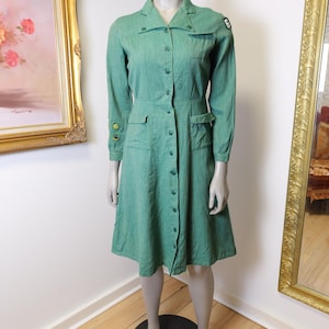 May include: A vintage green dress with long sleeves, a collared neckline, and a button-down front. The dress features two front pockets and a fitted waist. A patch with the number 6 is on the shoulder. The dress is displayed on a mannequin.