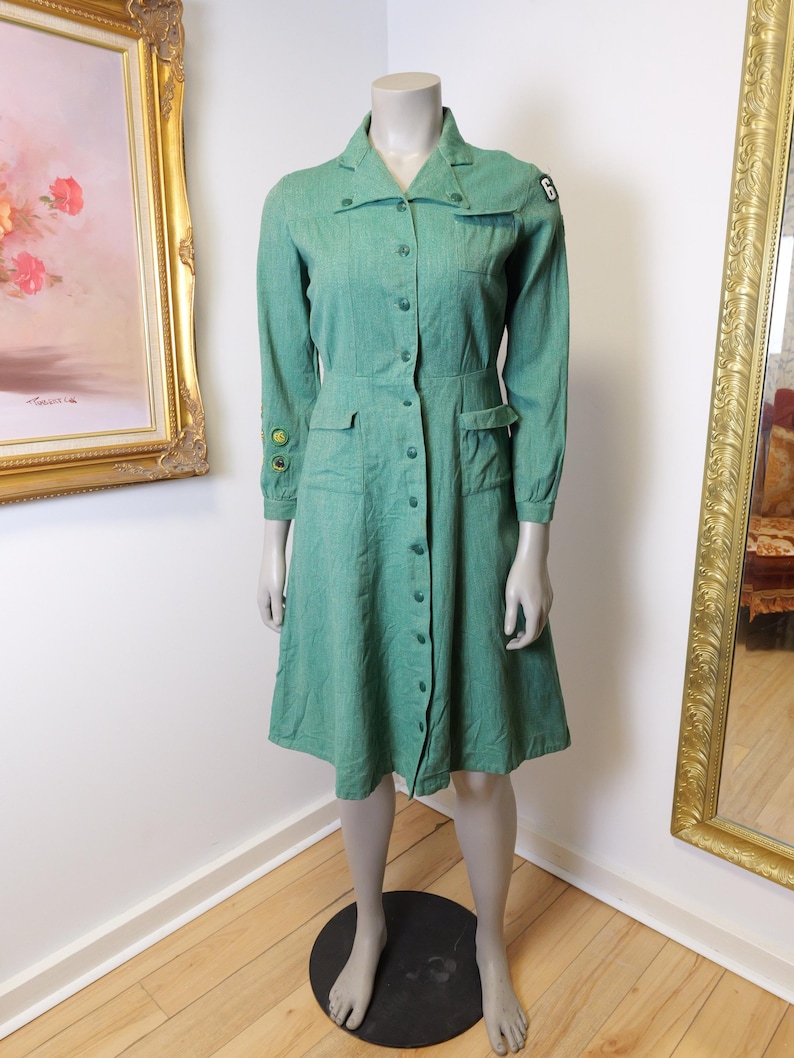 May include: A vintage green dress with long sleeves, a collared neckline, and a button-down front. The dress features two front pockets and a fitted waist. A patch with the number 6 is on the shoulder. The dress is displayed on a mannequin.
