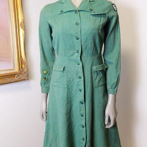 May include: A vintage green dress with long sleeves, a collared neckline, and button-down front. The dress features two front pockets, a fitted waist, and patches on the sleeves. The dress is displayed on a mannequin.