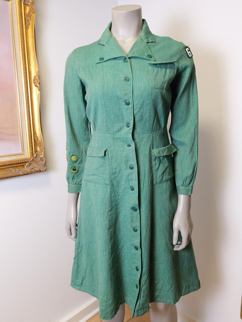 May include: A vintage green dress with long sleeves, a collared neckline, and button-down front. The dress features two front pockets, a fitted waist, and patches on the sleeves. The dress is displayed on a mannequin.