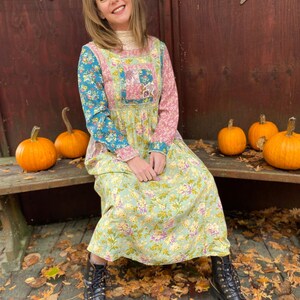 May include: A woman in a patchwork floral dress with long sleeves, seated on a wooden bench. The dress combines blue, pink, and green hues. She wears black boots. Several orange pumpkins are placed around her. The dress is a maxi length.
