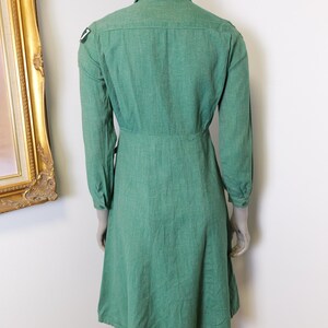 May include: A green long-sleeved dress with a collared neckline and a button-down front. The dress features a fitted waist and a flared skirt. A small patch is visible on the left shoulder. The dress is displayed on a mannequin.