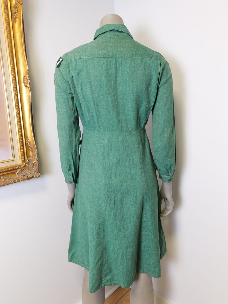 May include: A green long-sleeved dress with a collared neckline and a button-down front. The dress features a fitted waist and a flared skirt. A small patch is visible on the left shoulder. The dress is displayed on a mannequin.
