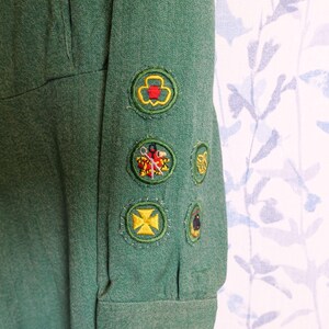 May include: Close-up of a green uniform sleeve with several embroidered patches. The patches feature various designs, including a trefoil, a cross, and other colorful emblems. The fabric has a textured appearance.