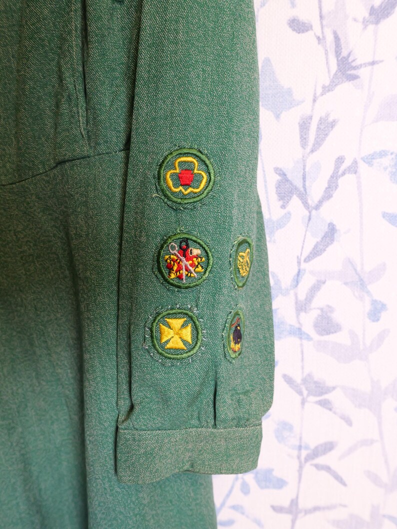 May include: Close-up of a green uniform sleeve with several embroidered patches. The patches feature various designs, including a trefoil, a cross, and other colorful emblems. The fabric has a textured appearance.