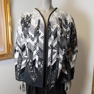 May include: A monochrome patchwork jacket with a striking design. The jacket showcases a mix of geometric and floral patterns in black and white, with black trim. The garment is displayed on a mannequin.