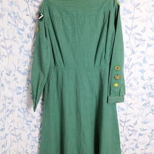 May include: A vintage green dress with long sleeves and a collared neckline. The dress features several patches on the sleeves and shoulder. The dress is hanging on a metal hanger against a floral patterned background.