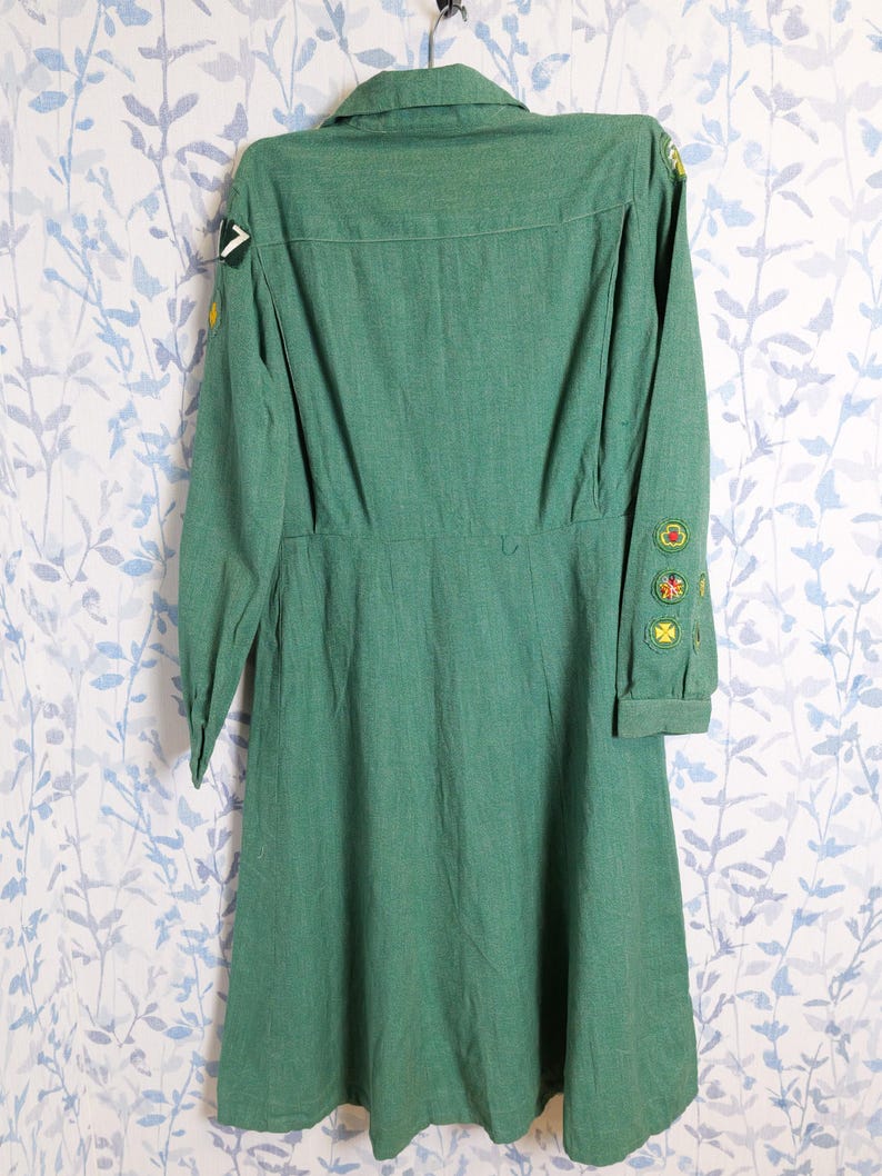 May include: A vintage green dress with long sleeves and a collared neckline. The dress features several patches on the sleeves and shoulder. The dress is hanging on a metal hanger against a floral patterned background.