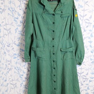May include: A vintage green dress with long sleeves, a collared neckline, and a button-down front. The dress features two front pockets and a fitted waist. A yellow clover patch is visible on the sleeve. The dress is hanging on a hanger.