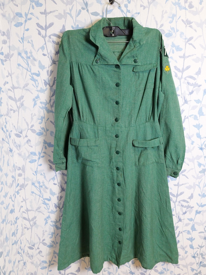 May include: A vintage green dress with long sleeves, a collared neckline, and a button-down front. The dress features two front pockets and a fitted waist. A yellow clover patch is visible on the sleeve. The dress is hanging on a hanger.