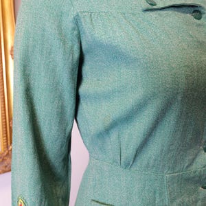 May include: A vintage, light green dress with long sleeves and a collared neckline. The dress features a fitted waist, a small pocket, and decorative embroidery on the sleeve. The fabric has a subtle textured appearance, and the dress is buttoned down the front.