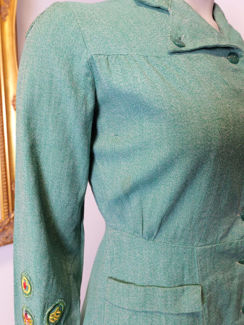 May include: A vintage, light green dress with long sleeves and a collared neckline. The dress features a fitted waist, a small pocket, and decorative embroidery on the sleeve. The fabric has a subtle textured appearance, and the dress is buttoned down the front.