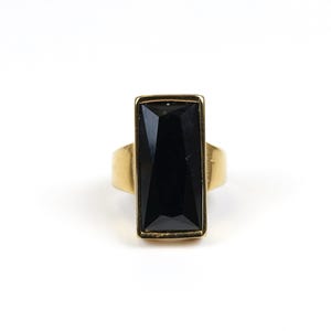 May include: A gold ring with a large, rectangular black gemstone. The gemstone is set in a gold frame, and the band has a simple, smooth design. The ring is shown against a white background.