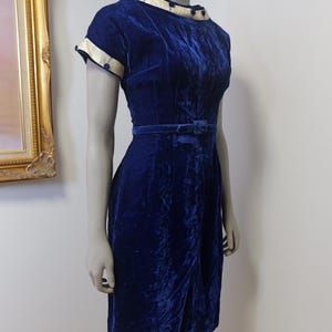Op de afbeelding: Een vintage blauwe fluwelen jurk met een witte kraag en manchetten. De jurk heeft een getailleerd bovenlijf en een volle rok. Het is versierd met kleine blauwe knopen.
