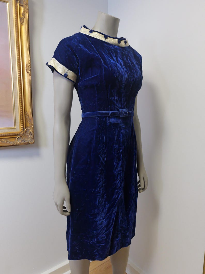 Op de afbeelding: Een vintage blauwe fluwelen jurk met een witte kraag en manchetten. De jurk heeft een getailleerd bovenlijf en een volle rok. Het is versierd met kleine blauwe knopen.