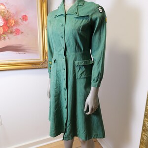 May include: A vintage green dress with long sleeves, a button-down front, and pockets. The dress features a collared neckline and a patch with the number 6 on the shoulder. The dress is displayed on a mannequin.