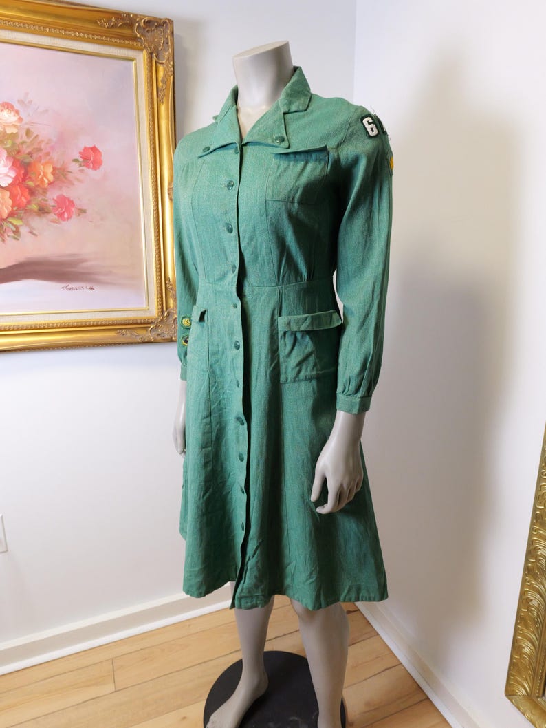 May include: A vintage green dress with long sleeves, a button-down front, and pockets. The dress features a collared neckline and a patch with the number 6 on the shoulder. The dress is displayed on a mannequin.