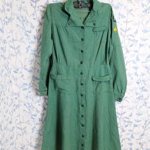 May include: A vintage green dress with long sleeves, a button-down front, and a collared neckline. The dress features two front pockets and a fitted waist. A yellow clover emblem is visible on the sleeve. The dress is hanging on a hanger against a floral patterned background.