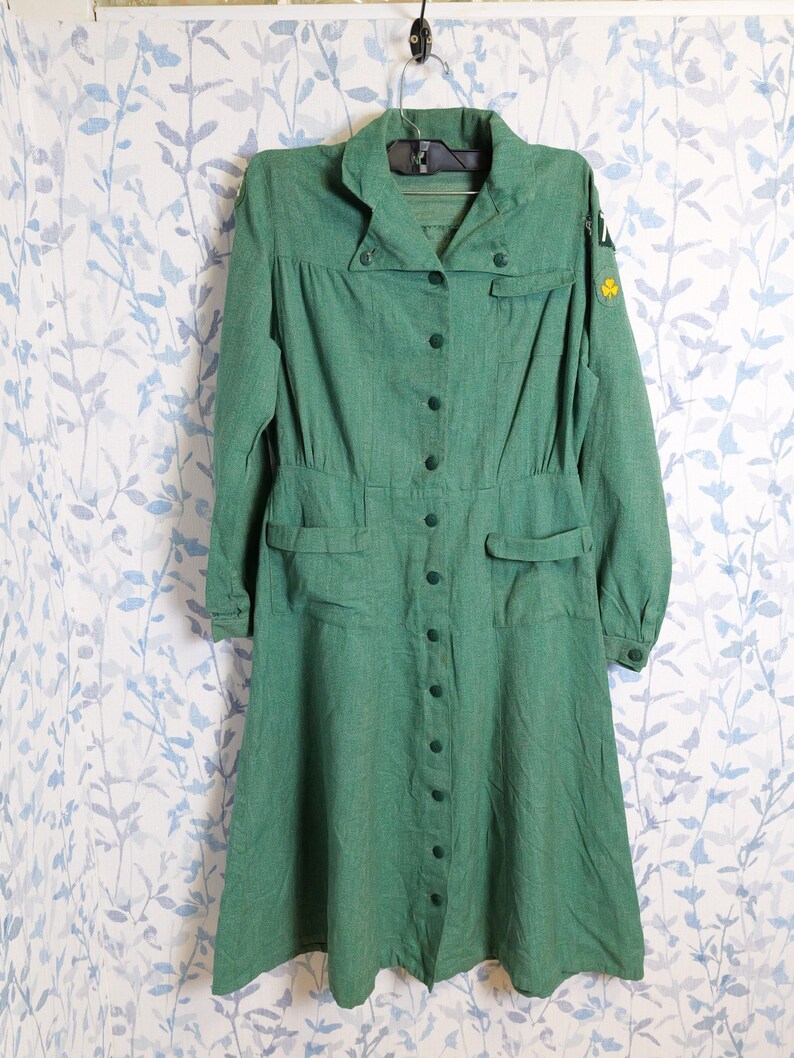 May include: A vintage green dress with long sleeves, a button-down front, and a collared neckline. The dress features two front pockets and a fitted waist. A yellow clover emblem is visible on the sleeve. The dress is hanging on a hanger against a floral patterned background.