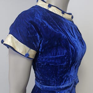 Op de afbeelding: Een koninklijk blauwe fluwelen jurk met een witte en gouden bies rond de halslijn en mouwen. De jurk heeft een getailleerd bovenlijf en een volle rok. Er zitten blauwe knopen op de halslijn en mouwen.