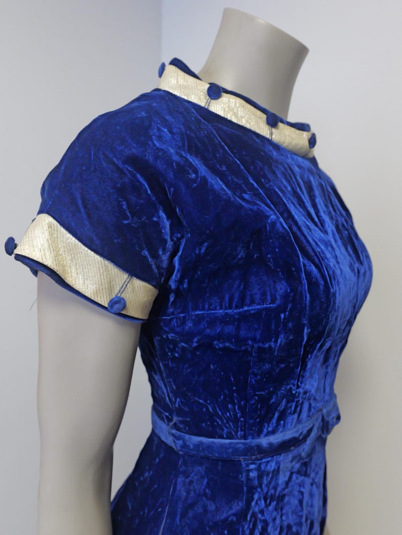 Op de afbeelding: Een koninklijk blauwe fluwelen jurk met een witte en gouden bies rond de halslijn en mouwen. De jurk heeft een getailleerd bovenlijf en een volle rok. Er zitten blauwe knopen op de halslijn en mouwen.