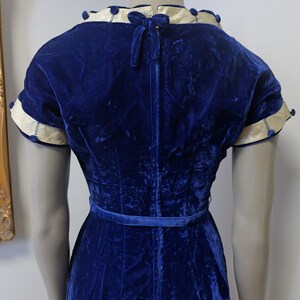 Op de afbeelding: Een koninklijk blauwe fluwelen jurk met een witte en blauwe bies rond de halslijn en mouwen. De jurk heeft een getailleerd bovenlijf en een volle rok. De jurk is in de taille getailleerd met een bijpassende blauwe riem.