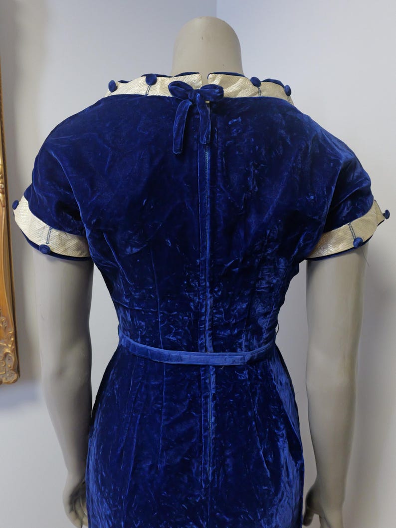 Op de afbeelding: Een koninklijk blauwe fluwelen jurk met een witte en blauwe bies rond de halslijn en mouwen. De jurk heeft een getailleerd bovenlijf en een volle rok. De jurk is in de taille getailleerd met een bijpassende blauwe riem.