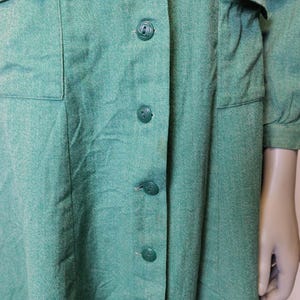 May include: A vintage green dress with a button-down front and two front pockets. The dress has long sleeves and a textured fabric. The buttons are a matching green color. The dress appears to be made of a woven material.