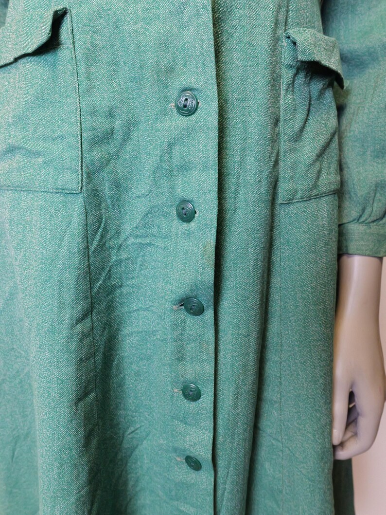 May include: A vintage green dress with a button-down front and two front pockets. The dress has long sleeves and a textured fabric. The buttons are a matching green color. The dress appears to be made of a woven material.