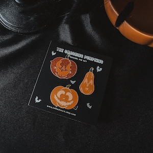 May include: A set of three enamel pins featuring orange and brown pumpkins with different facial expressions. The pins are on a black card with the text "The Smashing Pumpkins Enamel Pin Set" and a website address.