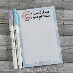 May include: A blue and white notepad with a donut illustration and the text "donut stress, you got this". Two pastel colored double-ended highlighters are next to the notepad.
