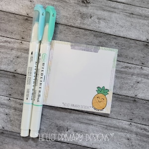 Op de afbeelding: Twee witte Zebra Mildliner markeerstiften met blauwe en groene doppen en een notitieblok met een lachende ananasafbeelding. Het notitieblok heeft de tekst "HELLO PRIMARY DESIGNS". De achtergrond is een houten oppervlak.