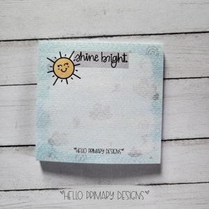 May include: A blue and white notepad with a smiling sun and the text "shine bright." The notepad has a textured surface and the text "HELLO PRIMARY DESIGNS" is printed on the bottom.