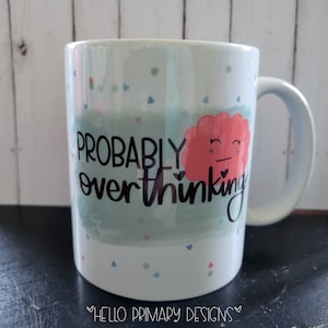 Puede incluir: Taza de cerámica blanca con un fondo acuarela azul claro y verde. La taza tiene una nube rosa con una cara y el texto "PROBABLY overthinking" en negro.