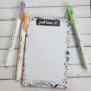 May include: A white notepad with a black and white doodle pattern border. The notepad has a white background and the text "just boo it!" written in black. Three colourful double-ended markers are lying on a white wooden surface next to the notepad. The markers are purple, orange, and green.