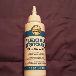 May include: A white bottle of Aleene's Flexible Stretchable Fabric Glue, with a blue and white label. The label reads "Flexible Stretchable Fabric Glue" and "4 fl oz (118 mL)". The glue is designed for t-shirts, knits, and stretch fabrics.