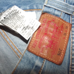 Pode incluir: Um close-up de uma etiqueta de jeans Levi's 501 em denim com o texto "Levi Strauss & Co. Original riveted Quality Clothing XX" e "501" em vermelho. A etiqueta também mostra o tamanho "W 36 L 34".
