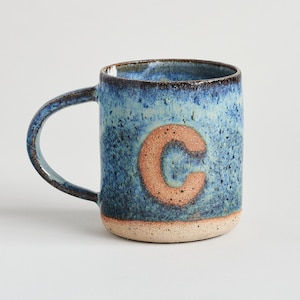 May include: A blue ceramic mug with a brown letter "C" on the front. The mug has a speckled glaze and a brown rim.
