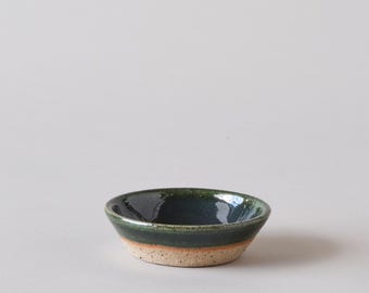 May include: A small, green ceramic bowl with a light brown rim. The bowl is glazed and has a slightly textured surface.