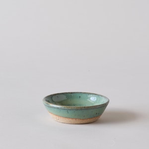 Könnte beinhalten: Eine kleine, hellgrüne Keramikschale mit einem gesprenkelten Überzug. Die Schale hat eine leicht abgerundete Form und einen leicht erhöhten Rand.