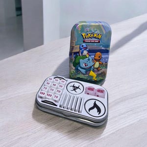 May include: A metal Pokemon trading card game tin featuring Bulbasaur, Squirtle, and Charmander. A white and silver game accessory with dice and number indicators is in front of the tin.