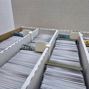 May include: A cardboard storage box with multiple compartments filled with stacks of cards. Each compartment has a white divider and a label with text. The labels read "PAR", "JTG", and "SVI".