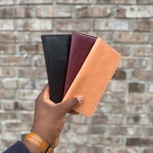 Op de afbeelding: Drie slanke, rechthoekige lederen etuis in zwart, donkerbordeaux en natuurlijk beige. Elk etui is voorzien van zichtbare witte stiksels langs de randen en heeft een gladde, matte afwerking. De items zijn in een waaier gerangschikt.