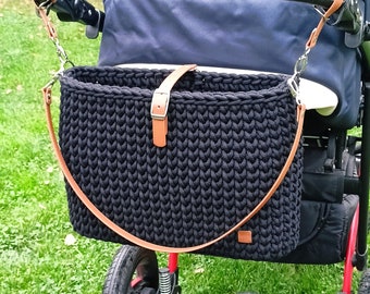 May include: A black crocheted rectangular bag with a tan leather strap and buckle closure, attached to the handle of a dark blue stroller. The bag features a thick, textured knit pattern and is suspended by two silver metal clips on the stroller handle.