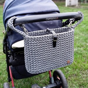 May include: A dark gray crocheted stroller organizer bag with a black leather strap and buckle closure. The bag features a braided gray cord handle attached to the black handlebar of a navy blue stroller with the brand name CAMARELO printed on it.