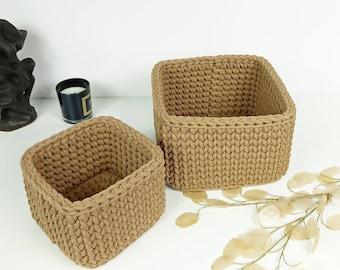 May include: Two square-shaped, brown, crocheted storage baskets. The baskets are made of a thick, textured yarn and have a woven pattern. The larger basket is in the background and the smaller basket is in the foreground.